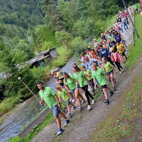 Eine Gruppe von Menschen in grünen Shirts läuft auf einem Pfad neben einem Fluss. Sie scheinen Teil einer geführten Tour zu sein, angeführt von einem Mann in einem grünen Shirt, der einen Stock hält.