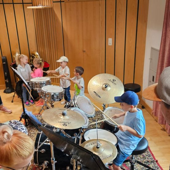 Mehrere Kinder spielen Trommeln und Becken in einem Musikraum, wobei ein Junge in einer blauen Kappe anführt.