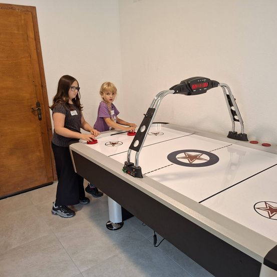 Zwei junge Mädchen spielen Air Hockey in einem Raum mit einer Holztür. Der Tisch hat eine schwarz-weiße Spielfläche mit Sternen.