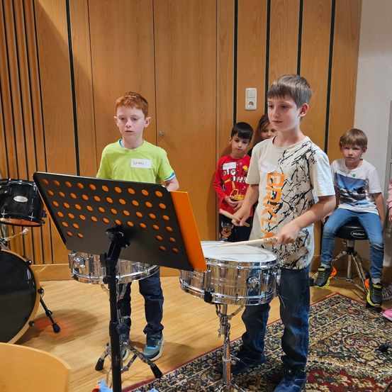 Zwei junge Jungen spielen Trommeln in einem Musikraum, mit einem Notenständer und Notenblättern vor ihnen. Andere Kinder beobachten sie dahinter.