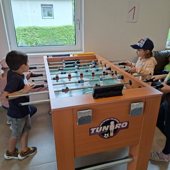 Zwei Kinder spielen Tischfußball in einem Raum. Der Junge auf der linken Seite schaut auf den Tisch. Das Mädchen auf der rechten Seite trägt eine Mütze.