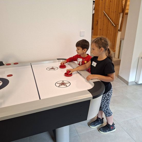 Zwei Kinder, ein Junge und ein Mädchen, spielen Air Hockey drinnen. Der Junge trägt ein rotes Shirt, während das Mädchen ein schwarzes Shirt trägt. Sie stehen vor einem Tisch mit roten Pucks.