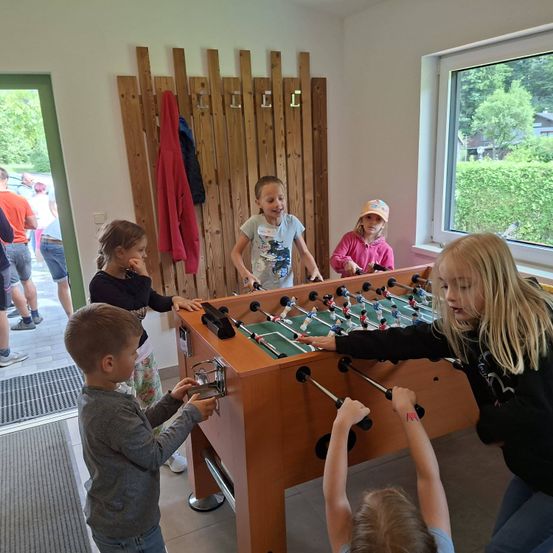 In einem Raum spielen eine Gruppe von Kindern Tischfußball. Sie stehen um einen Holztisch mit Griffen herum, während zwei Kinder Armbänder tragen. Im Hintergrund befinden sich eine Tür und ein Fenster.