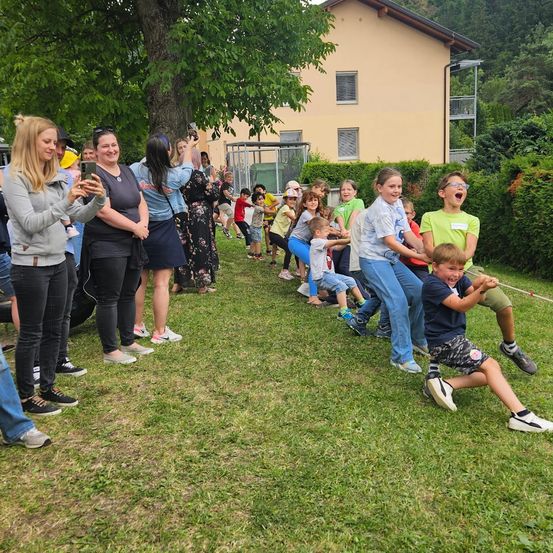 Gruppe von Erwachsenen und Kindern nimmt an einem Tauziehen auf einem Rasenfeld teil, während Zuschauer den Moment festhalten. Ein Haus mit Balkonen ist im Hintergrund.