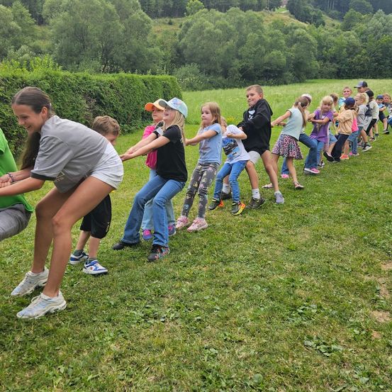 Eine Gruppe von Kindern und Erwachsenen spielt eine Partie Tauziehen auf einem Grasfeld. Sie sind in zwei Teams aufgeteilt.