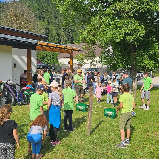 Eine Gruppe von Menschen versammelt sich auf einer Rasenfläche. Einige tragen grüne T-Shirts, andere sind in Freizeitkleidung. Sie stehen im Kreis um ein Seil. Im Hintergrund befinden sich Zelte und Autos.