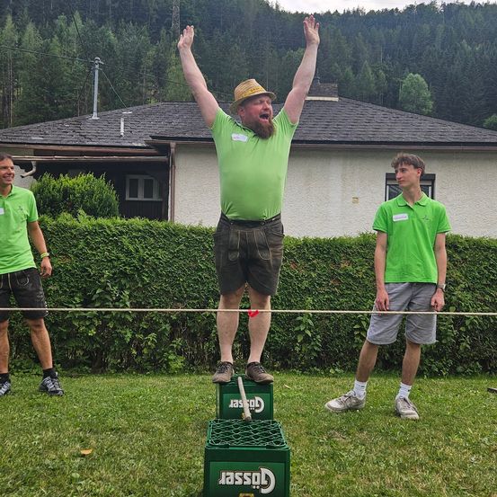 Drei Männer in grünen Shirts stehen auf einem Rasen. Einer balanciert auf einem Kistenstapel und hält eine Schnur, während zwei andere zusehen. Ein Haus mit einem schiefergedeckten Dach und Grünpflanzen steht dahinter.