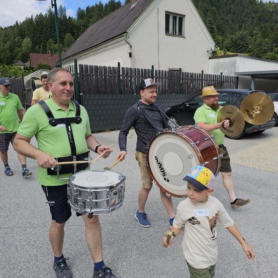 Eine Gruppe von Männern in grünen Hemden spielt Trommeln im Freien. Hinter ihnen hält ein kleiner Junge einen Drumstick und beobachtet. Sie sind umgeben von Häusern, Bäumen und einem geparkten Auto.
