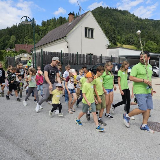 Eine Gruppe von Kindern und Erwachsenen in grünen Shirts läuft auf einer Straße, mit zwei Männern, die vorne führen. Dahinter steht ein Haus mit einem Zaun und einer Straßenlaterne.