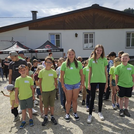 Eine Gruppe von Kindern in grünen T-Shirts posiert für ein Foto vor einem Gebäude mit 24MX-Schildern. Ein Kind hält einen Hammer.