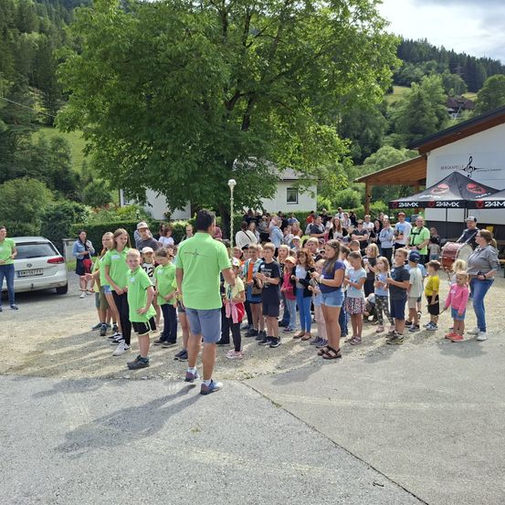 Eine Menschenmenge, einschließlich Kinder in grünen Shirts, versammelt sich draußen in der Nähe eines Gebäudes. Bäume und Berge sind im Hintergrund.