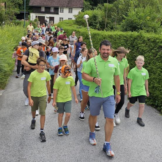 Eine Gruppe von Menschen in grünen T-Shirts geht einen Pfad entlang. Sie werden von einem Mann mit einem Stock angeführt. Bäume und Büsche säumen die Seiten des Pfades. Im Hintergrund steht ein Haus mit einem braunen Dach.