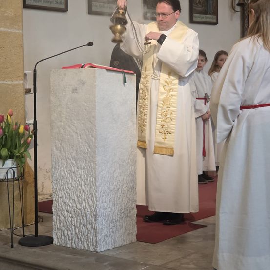 Ein Priester steht an einer Kanzel, hält eine Glocke und zwei Frauen in weißen Gewändern beobachten ihn. Ein Mikrofon und eine Pflanze sind in der Nähe.