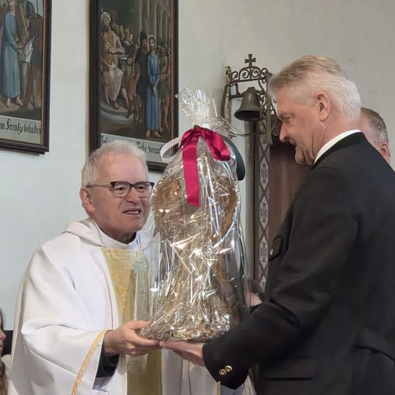 Ein Priester in einer weißen Robe überreicht einem Mann in formeller Kleidung ein verpacktes Geschenk in einer Kirche.