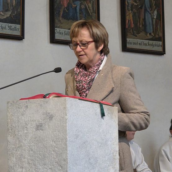 Eine ältere Frau steht an einem Pult mit Mikrofon und Buch und liest, während andere zusehen. Drei gerahmte Gemälde hängen an der Wand hinter ihr.