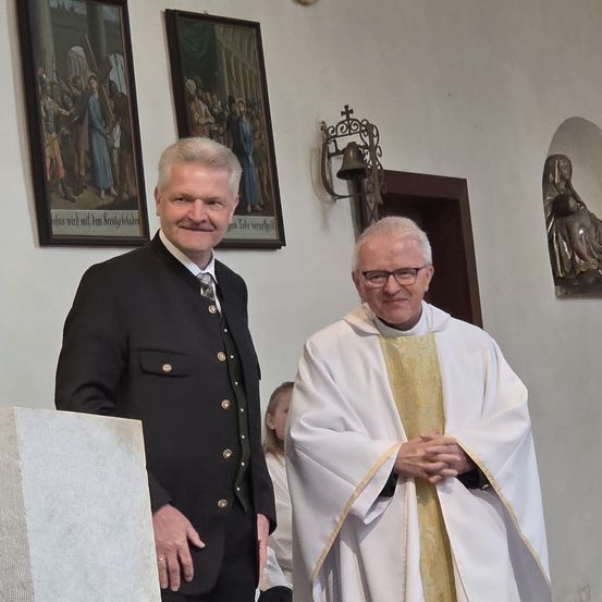 Zwei Männer stehen in einer Kirche, einer in formeller Kleidung, der andere in einem Priestergewand. Hinter ihnen hängen zwei Gemälde und eine Glocke an der Wand.