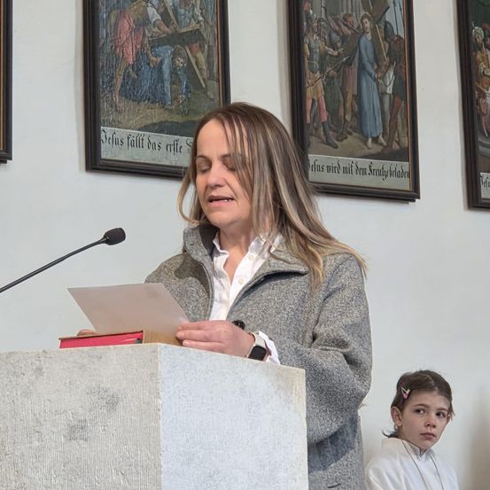 Eine Frau spricht an einem Podium mit Mikrofon. Sie hält ein Papier in der Hand und sieht ernst aus. Hinter ihr hängen drei gerahmte Bilder an der Wand. Ein junges Mädchen schaut von der Seite.