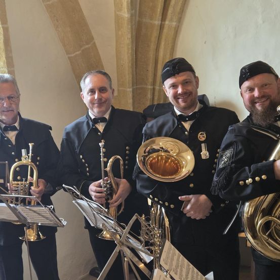 Vier Männer in schwarzen Uniformen mit Medaillen stehen in einem Raum und halten Trompeten und Tuben, lächeln für ein Foto.