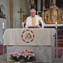 Ein Priester steht am Altar und hält eine Predigt. Kerzen und ein Kreuz schmücken den Altar, und Blumenarrangements sind davor platziert. Hinter ihm steht eine Statue eines Heiligen und ein goldenes Altarbild.