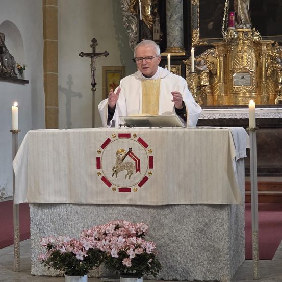 Ein Priester steht am Altar und hält eine Predigt. Kerzen und ein Kreuz schmücken den Altar, und Blumenarrangements sind davor platziert. Hinter ihm steht eine Statue eines Heiligen und ein goldenes Altarbild.