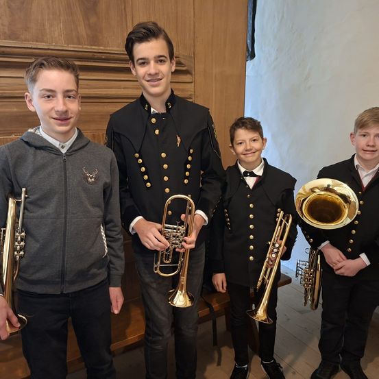Vier junge Männer stehen eng zusammen und halten Blechblasinstrumente. Der Mann auf der linken Seite trägt eine graue Kapuzenjacke, während die anderen schwarze Uniformen mit goldenen Knöpfen tragen.