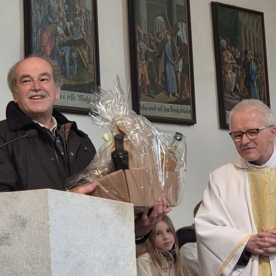 Ein Mann mit einem Mikrofon an einem Podium hält einen Korb mit Weinflaschen. Neben ihm steht ein Priester, und hinter ihnen hängen drei Gemälde an der Wand.