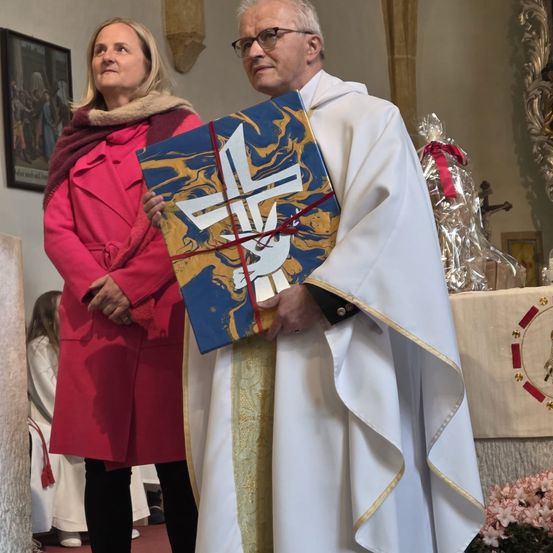 Ein Priester in einer weißen Robe, der ein verpacktes Buch in der Hand hält, steht mit einer Frau in einem rosa Mantel in einer Kirche.