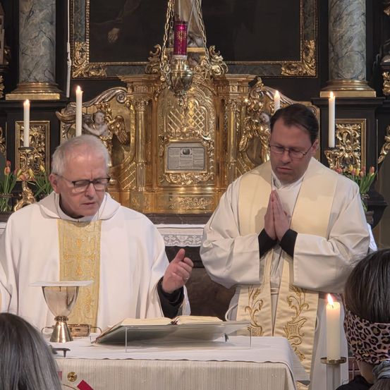Zwei Priester führen einen Gottesdienst in einer Kirche mit kunstvollem Altar und Kerzen durch. Einer spricht, und der andere betet mit gefalteten Händen.
