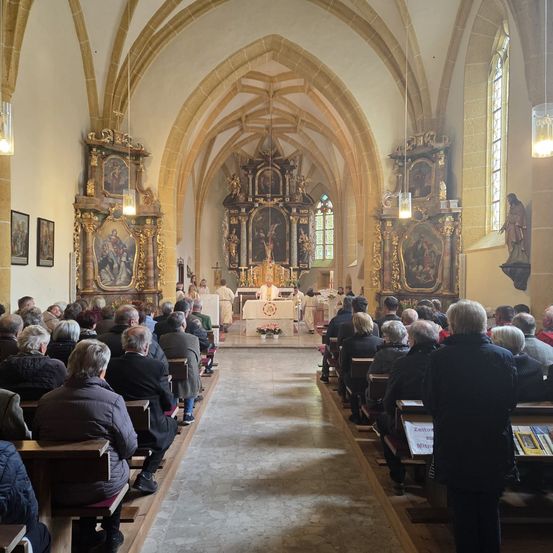 In einer Kirche sitzen die Besucher in Reihen, der Altar gegenüberstehend, wo Priester stehen. Der Altar ist mit Blumen und Kerzen geschmückt. Kunstwerke und Statuen schmücken die Wände.