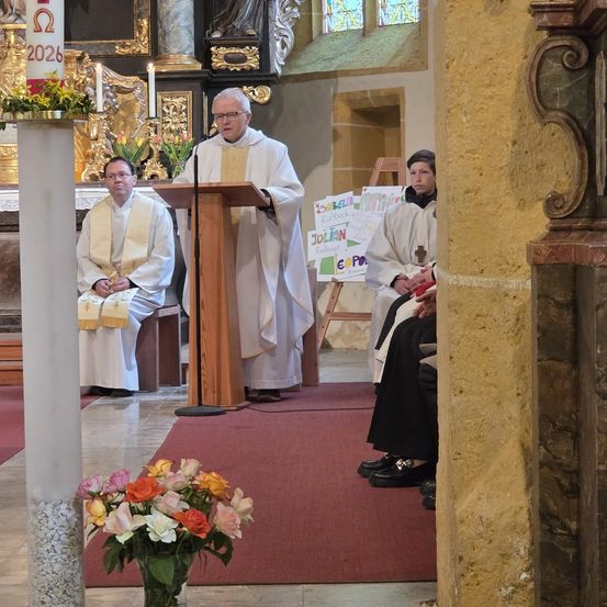 Ein Priester spricht an einer Kanzel in einer Kirche. Zwei weitere Priester sitzen hinter ihm. Ein roter Teppich verläuft durch die Mitte.
