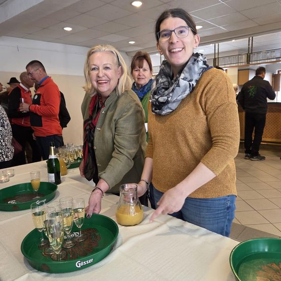 Zwei Frauen stehen hinter einem Tisch mit Champagner und Saft. Sie lächeln und unterhalten sich. Hinter ihnen stehen und reden Leute.