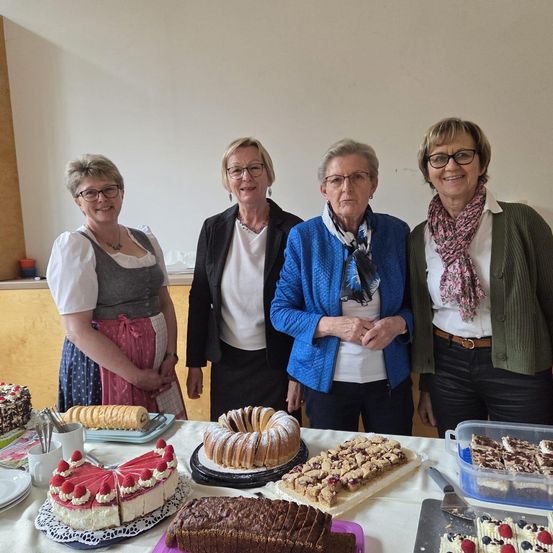 Vier Frauen stehen hinter einem Tisch voller Desserts. Die Frau auf der linken Seite trägt ein traditionelles deutsches Kleid. Die anderen tragen Brillen und Jacken. Auf dem Tisch befinden sich Kuchen, Brot und andere Backwaren.