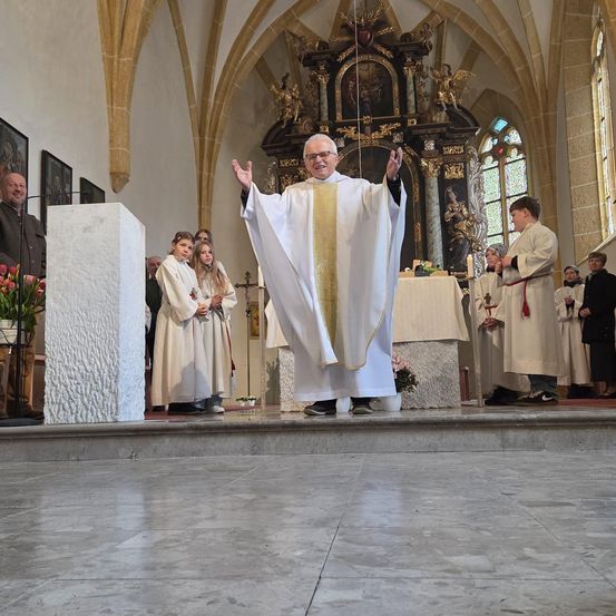 Ein Priester hält im Inneren einer Kirche eine Predigt. Er steht mit erhobenen Händen auf einer Bühne. Hinter ihm steht ein Pult, und links sind Blumen zu sehen.