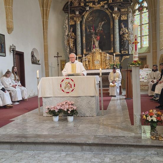 Ein Priester steht am Altar in einer Kirche, mit Kerzen und Blumen. Akolythen sitzen an den Seiten. Dahinter befindet sich ein Gemälde einer religiösen Szene.