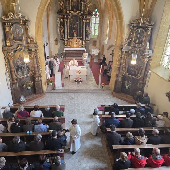 Ein Priester steht am Altar in einer Kirche mit auf den Bänken sitzenden Menschen. Der Altar hat Kerzen, Blumen und einen Tisch. Zwei Personen stehen in der Nähe des Altars. Die Kirche ist mit aufwendigen Verzierungen geschmückt.