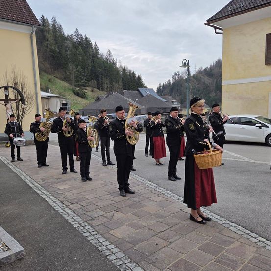 Eine Blaskapelle in schwarz-roten Uniformen steht auf einem gepflasterten Gehweg mit Bergen im Hintergrund.