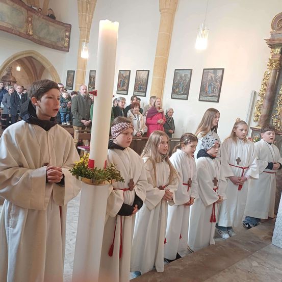 Kinder in weißen Gewändern halten eine Kerze während einer religiösen Zeremonie in einer Kirche mit gerahmten Bildern an der Wand und Menschen im Hintergrund.