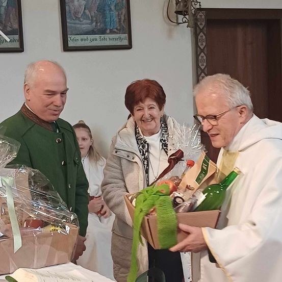 Ein Priester in einer weißen Robe hält eine Geschenkbox mit einem lächelnden Paar. Die Frau trägt eine Halskette und der Mann einen grünen Mantel. Dahinter steht ein Mädchen und schaut auf die Box.