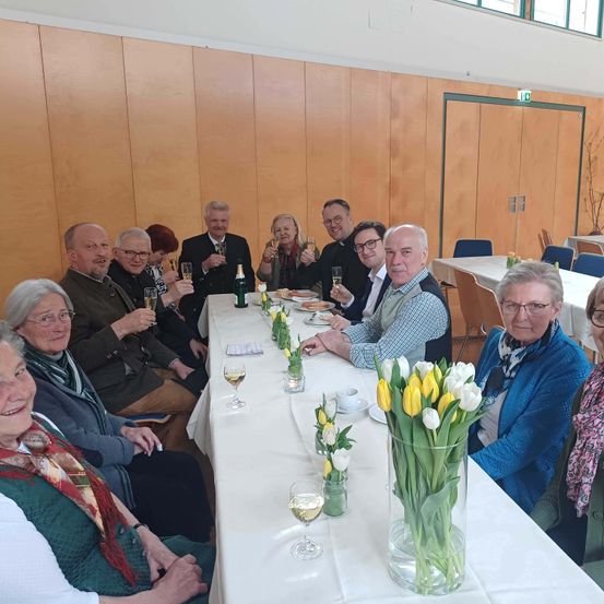Eine Gruppe Erwachsener sitzt um einen Tisch herum und prostet mit Weinläsern. Auf dem Tisch stehen Blumen. Ein Mann im Anzug hält eine Flasche. Sie sind in einem Raum mit Holzwänden.