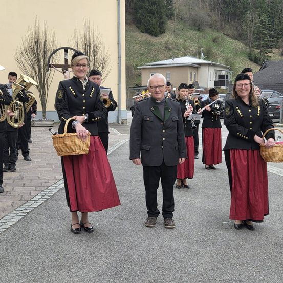 Eine Gruppe von Musikern in traditioneller Kleidung mit Körben steht vor einem Gebäude, ein Mann in einem Anzug in der Mitte. Im Hintergrund befinden sich Bäume, ein Kreuz und ein Gebäude.
