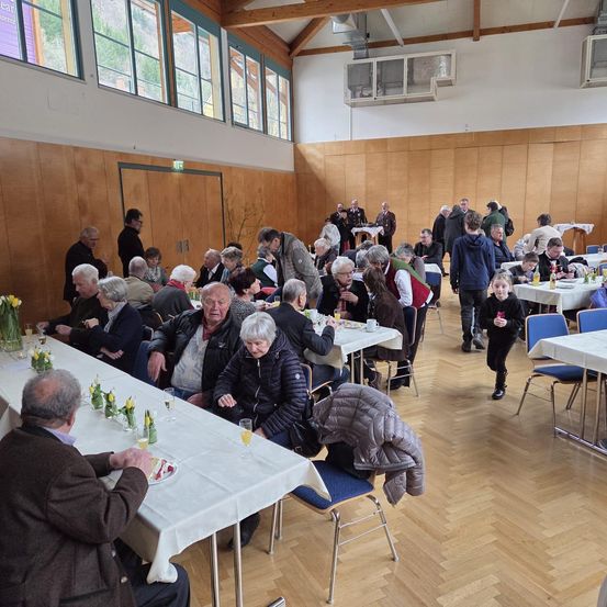 Eine Gruppe von Menschen sitzt um Tische in einem großen Raum mit Holzböden und großen Fenstern. Einige Leute stehen und reden, während andere essen oder trinken.