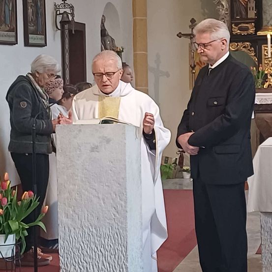 Ein Priester liest in einer Kirche von einem Buch am Kanzelpult, während ein anderer Mann neben ihm steht. Eine ältere Frau beobachtet in der Nähe, mit Blumen und einem Kreuz im Hintergrund.