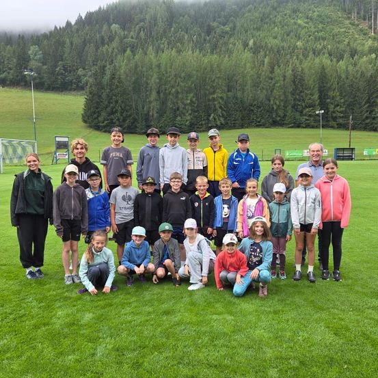Eine Gruppe von Kindern und Erwachsenen posiert für ein Foto auf einem Rasenfeld mit Bergen im Hintergrund.