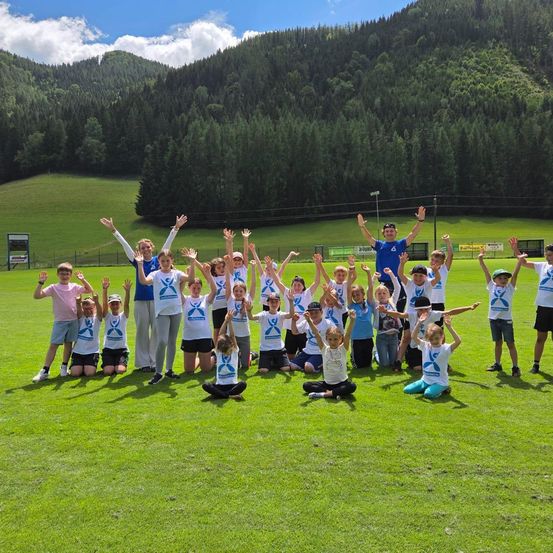 Eine Gruppe von Kindern und zwei Erwachsenen posiert für ein Foto auf einem Rasenfeld mit einem Hintergrund aus Bergen und Bäumen.
