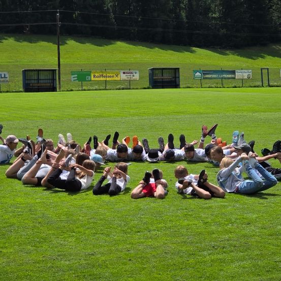 Eine Gruppe von Kindern liegt auf einem Grasfeld und bildet einen Kreis, wobei sich ihre Füße berühren. Sie tragen Turnschuhe und einige Mützen. Hinter ihnen befinden sich Werbebanner und ein Holzpfahl.