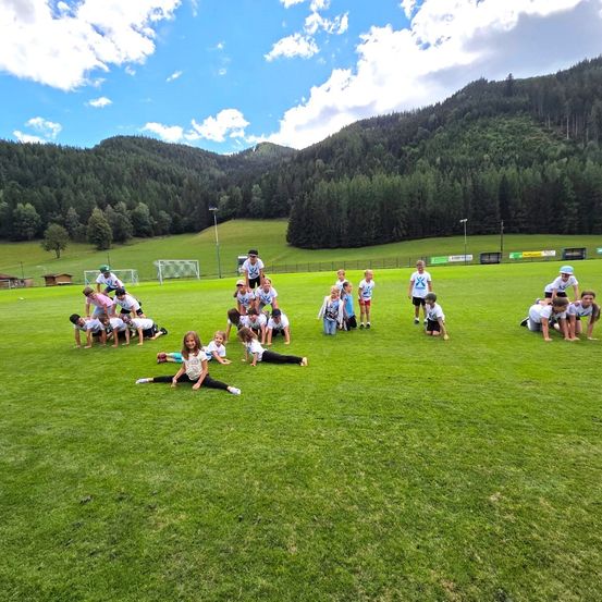 Eine Gruppe von Kindern spielt auf einem Rasenfeld mit Bergen im Hintergrund. Einige liegen, andere stehen oder knien. Sie tragen weiße Hemden und Turnschuhe.
