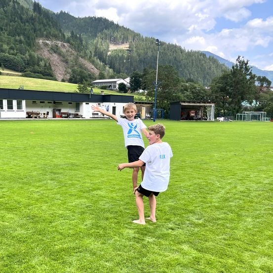 Zwei Jungen spielen auf einem Rasenfeld mit Bergen im Hintergrund. Ein Junge steht und lächelt, während der andere Junge springt.