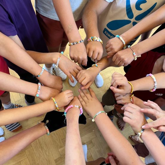 Eine Gruppe von Kindern und Jugendlichen hält sich an den Händen in einem Kreis, jeder trägt bunte Perlenarmbänder. Sie stehen auf einem Holzboden, wahrscheinlich in einer Schule oder einem Gemeindezentrum.
