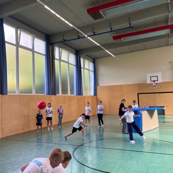 Eine Gruppe von Kindern spielt mit einem roten Ball in einer Sporthalle. Einige laufen, während andere stehen und zuschauen.