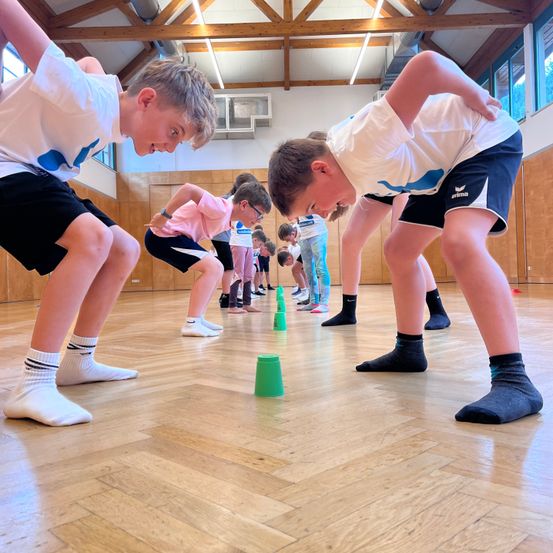 Eine Gruppe von Kindern in einer Turnhalle, die an einer körperlichen Aktivität teilnehmen. Sie stehen in einer Reihe, mit Blick nach vorne, mit grünen Bechern auf dem Boden. Die Kinder tragen weiße Socken und schwarze Shorts.